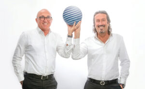 Rastelli e Bizzarri, fondatori del Polo Informatico, posano sorridenti in camicia bianca reggendo insieme una sfera a righe blu e bianche su sfondo bianco