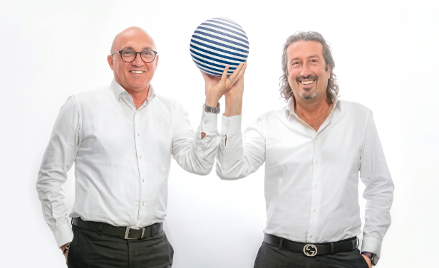 Rastelli e Bizzarri, fondatori del Polo Informatico, posano sorridenti in camicia bianca reggendo insieme una sfera a righe blu e bianche su sfondo bianco